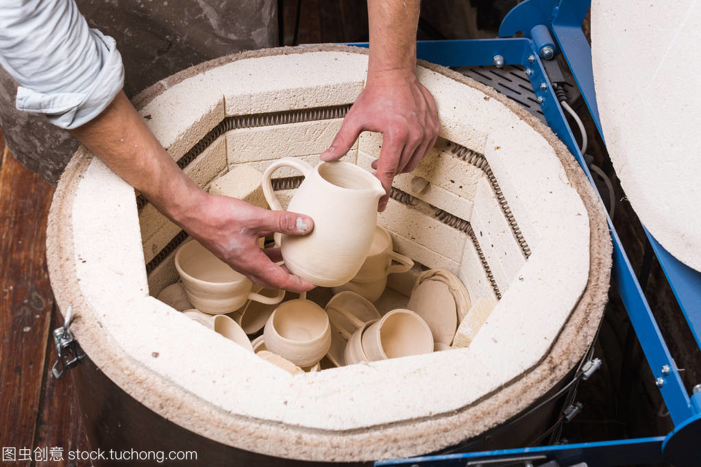 陶器、 车间、 陶瓷艺术概念-特写男性手上的轻轻地把壶窑,为进一步焙烧的未完成的粘土制品、 杯子和生的器具电烘箱的顶视图